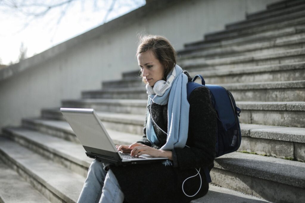 "A student sits outdoors on concrete steps while typing on her laptop computer. Furthermore, she utilises headphones to focus deeply during her comprehensive KS4/Study Skills online tutoring training session."