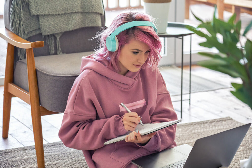 A young person with bright pink hair and mint-green headphones sitting on a rug on the floor, writing on a tablet with a stylus while looking down at an open laptop.