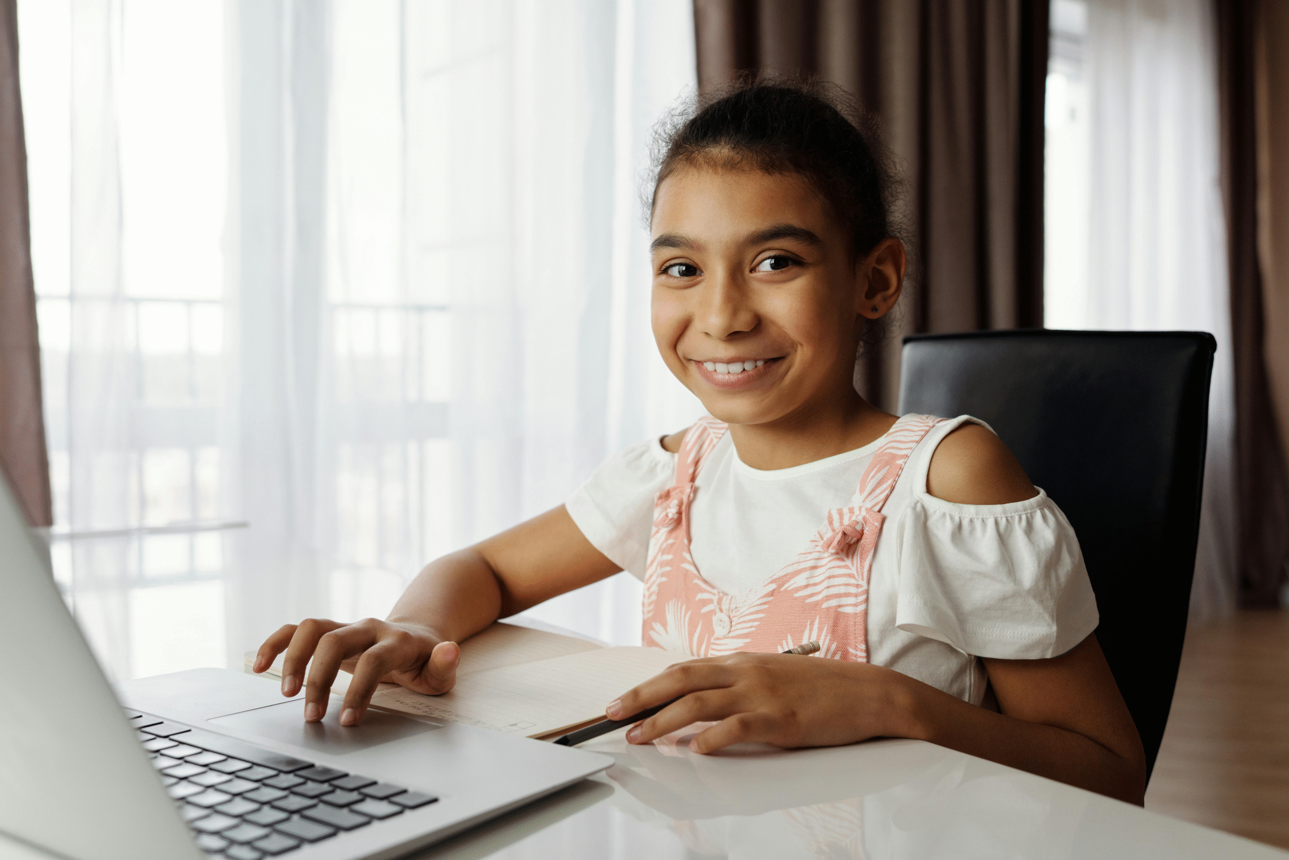 A young student smiling and engaged in an interactive online tutoring session with a laptop and notebook at home.