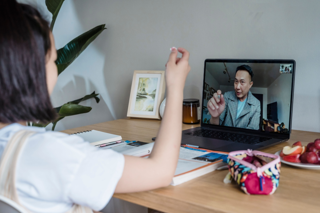 Over-the-shoulder view of a young student sitting at a wooden desk with open textbooks, holding up a small object during a video call with a tutor on a laptop.