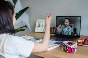 Over-the-shoulder view of a young student sitting at a wooden desk with open textbooks, holding up a small object during a video call with a tutor on a laptop.